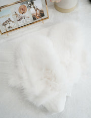 A white fur glove displayed on a flat surface, with a blurred background featuring a flower and a picture frame.