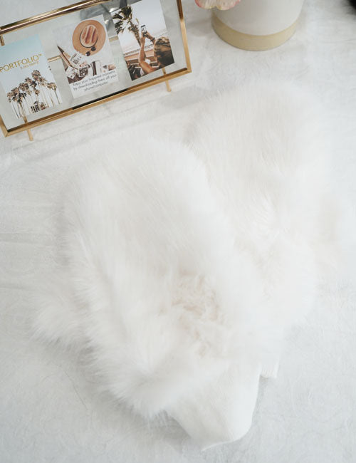 A white fur glove displayed on a flat surface, with a blurred background featuring a flower and a picture frame.